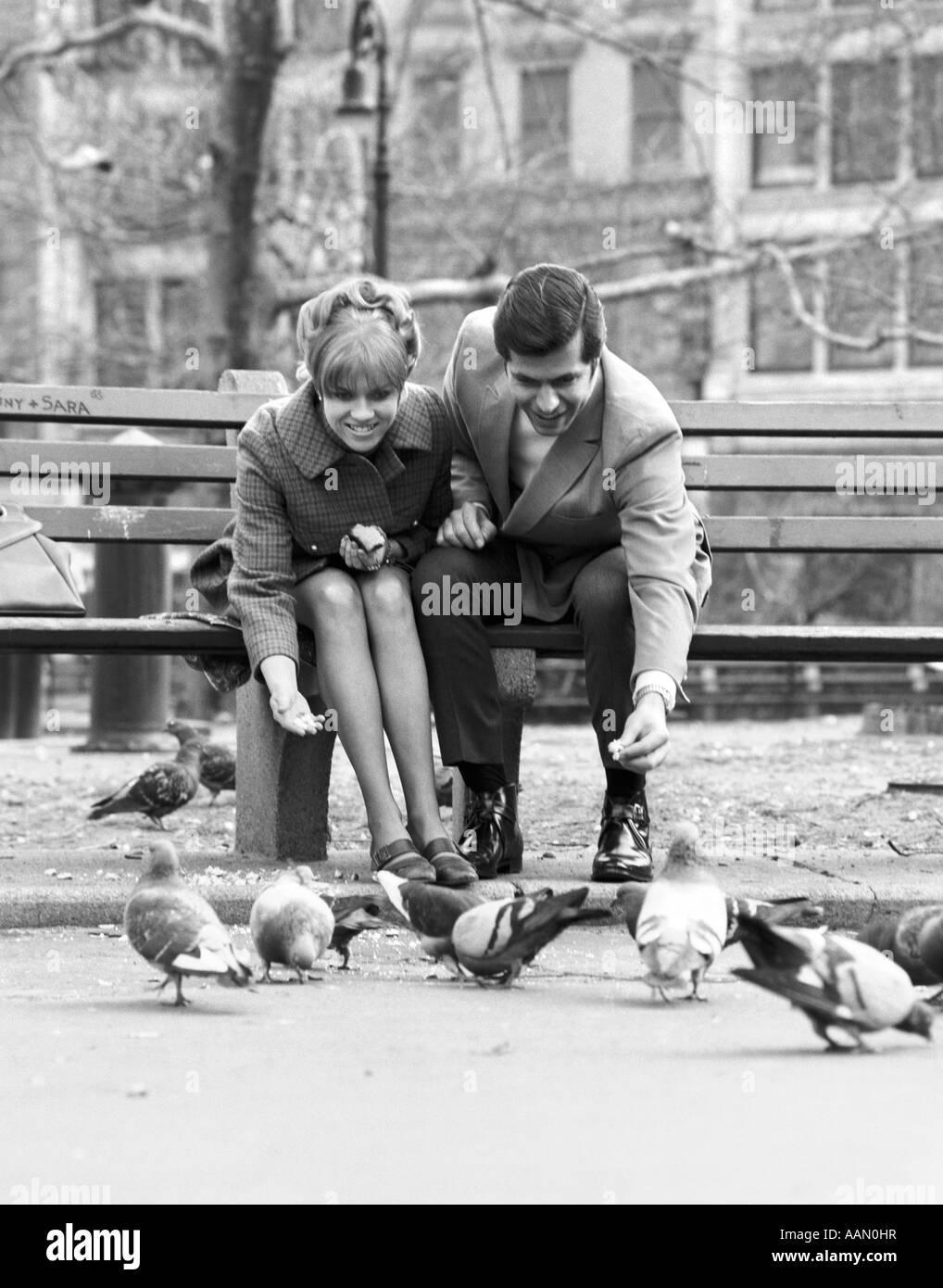 1970s COUPLE ON PARK BENCH LEANING DOWN TO FEED PIGEONS Stock Photo