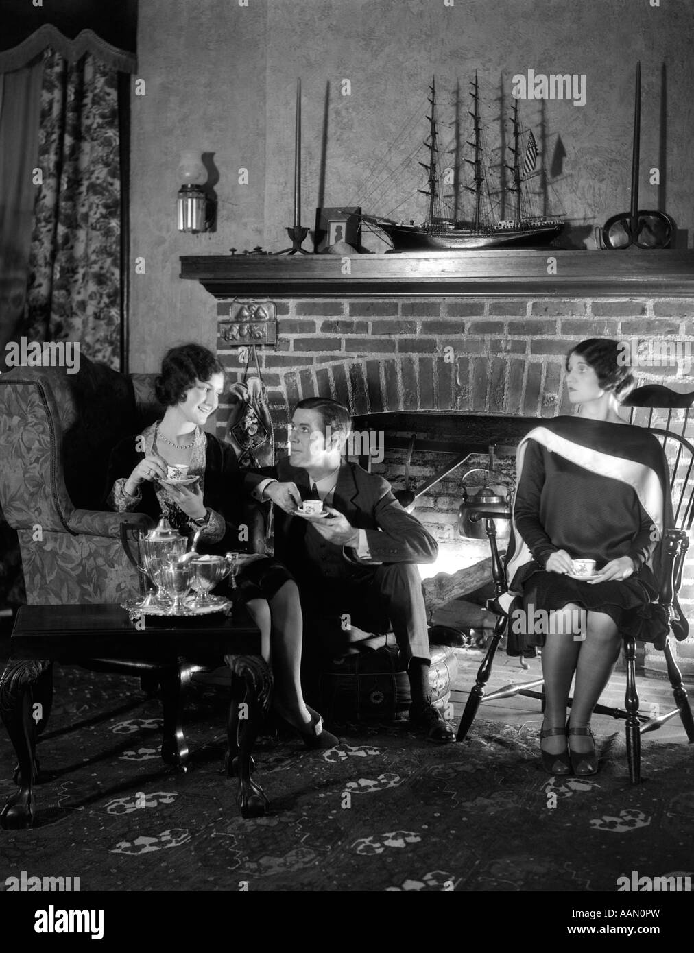 1920s MAN SITTING BETWEEN TWO WOMEN FLIRTING WITH ONE WOMAN IGNORING THE OTHER DRINKING TEA FIREPLACE IN BACKGROUND Stock Photo