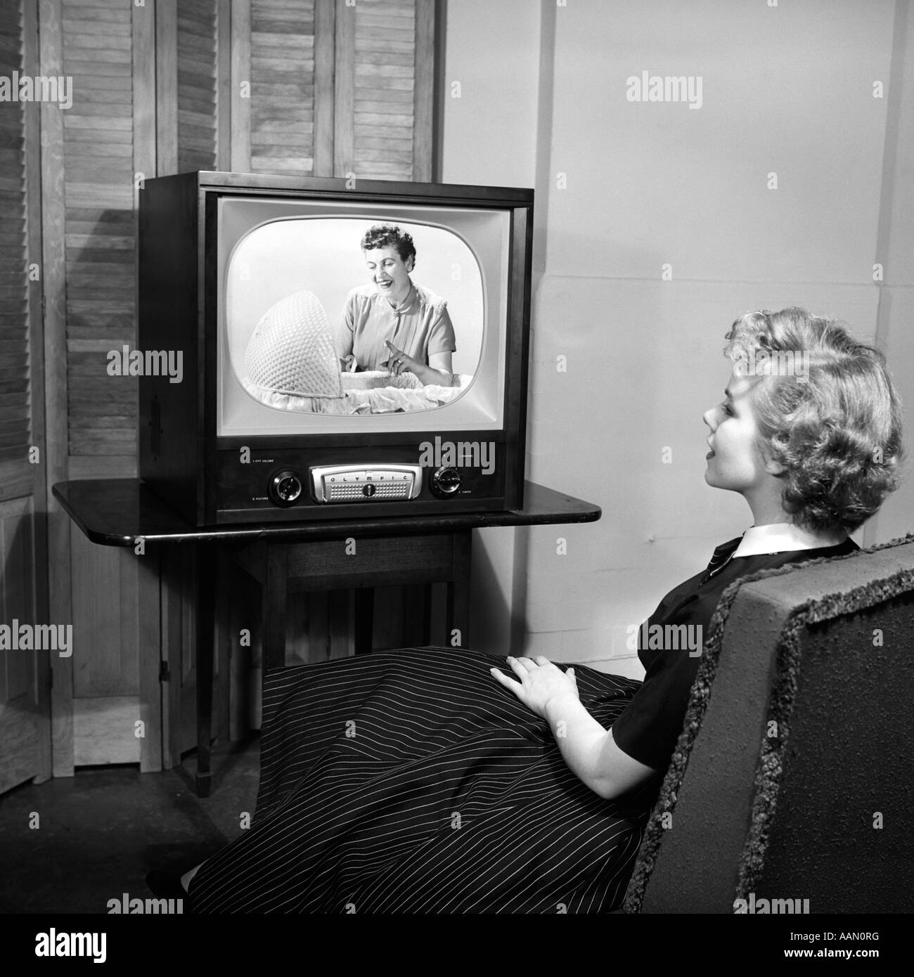 1950s TEEN SITTING WITH BACK TO VIEWER WATCHING A WOMAN LOOKING INTO A BASSINET ON HER TELEVISION IN A DARK ROOM Stock Photo