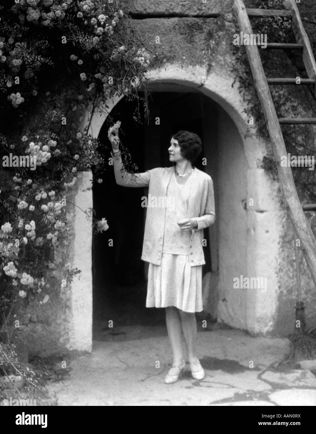 1930s WOMAN IN FLAPPER OUTFIT STANDING IN FRONT OF WHITEWASHED ARCHWAY WITH FLOWERS PICKING BUD BRITTANY FRANCE Stock Photo