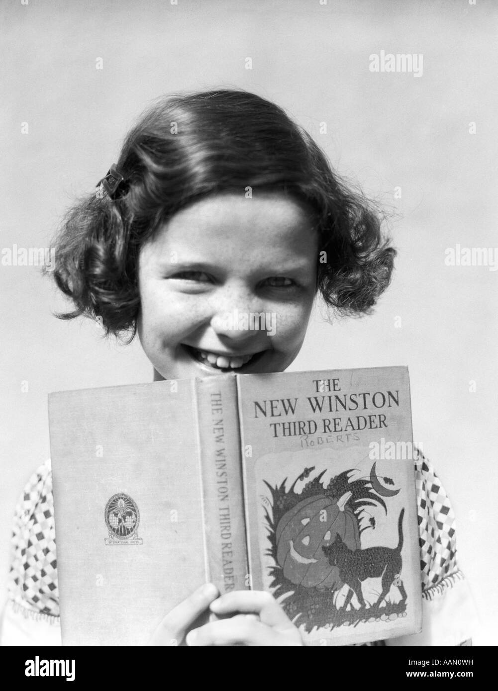 1930s SMILING GIRL WITH BOBBED HAIR HOLDING AN OPEN THIRD GRADE READING BOOK Stock Photo