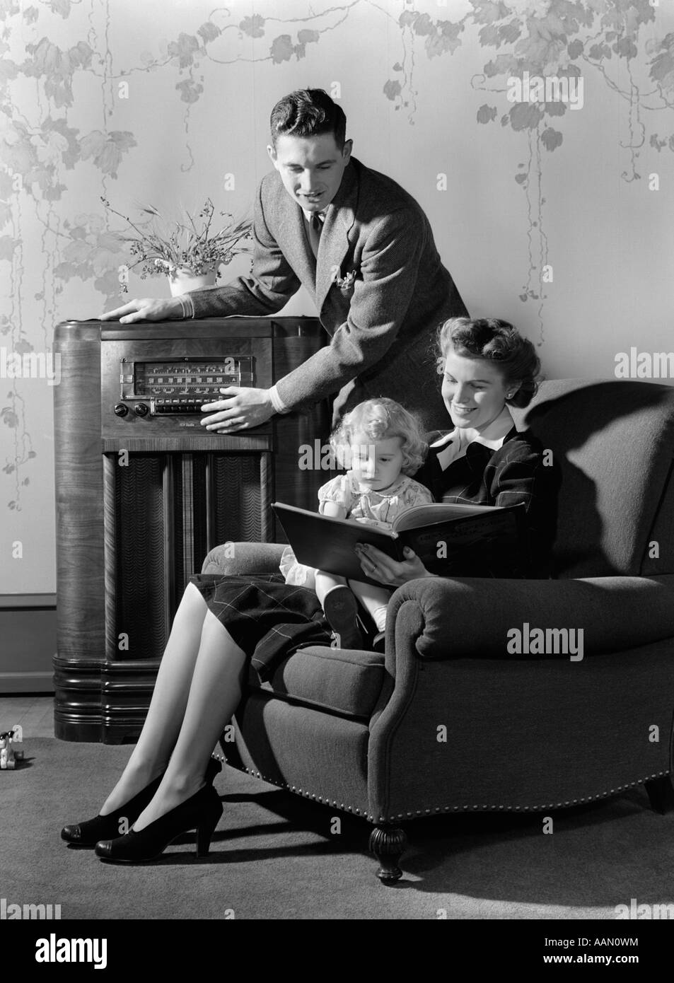 1930s 1940s FAMILY LISTENING TO RADIO WHILE MOTHER READS BOOK TO LITTLE GIRL SEATED IN HER LAP IN ARMCHAIR Stock Photo