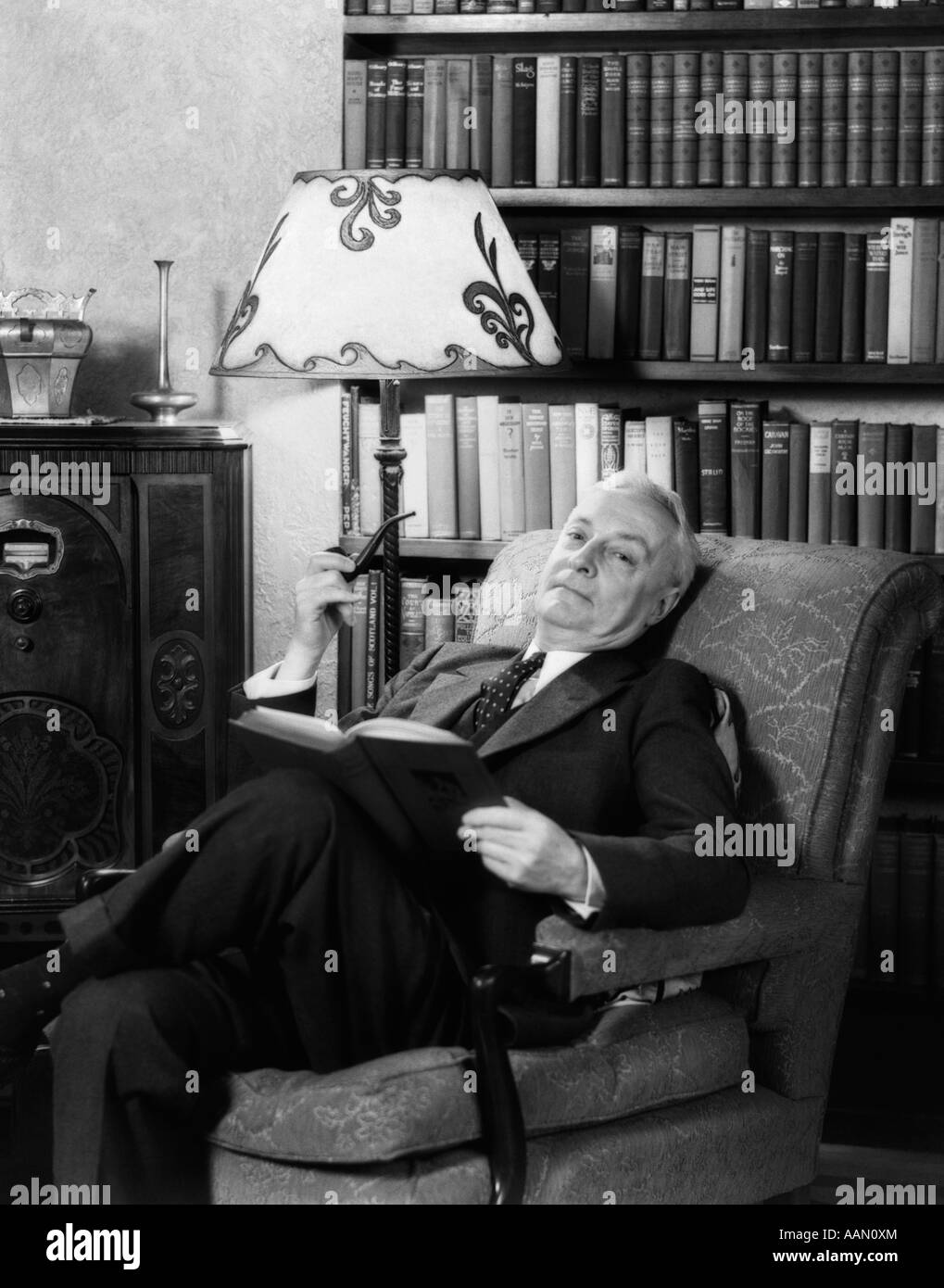 1930s ELDERLY MAN IN ARMCHAIR RELAXING WITH BOOK & PIPE & LISTENING TO RADIO Stock Photo