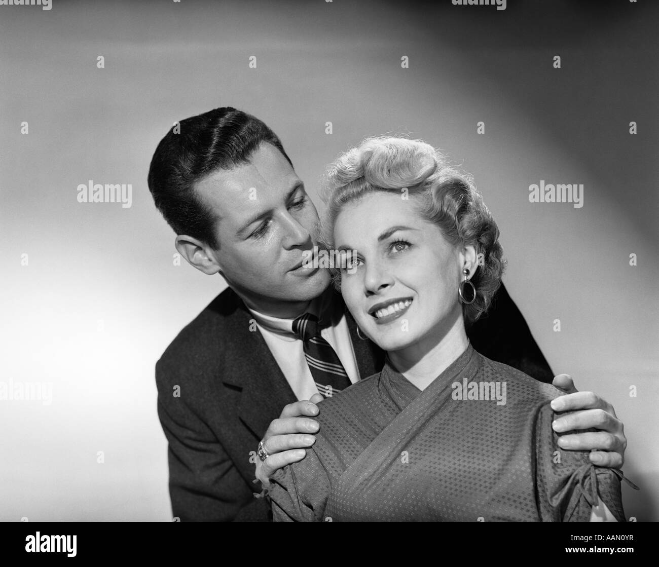1950s COUPLE PORTRAIT SMILING MAN BEHIND WOMAN WHISPER IN EAR HANDS ON HER SHOULDERS Stock Photo