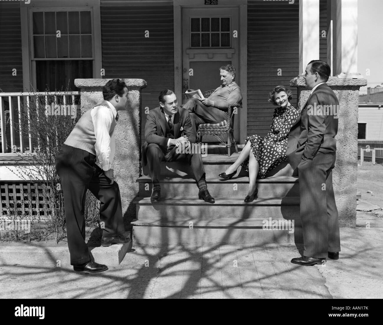 1940s GROUP OF FIVE MEN & WOMEN GATHERED ON & AROUND FRONT PORCH FOUR IN FRONT TALKING ELDERLY MAN IN BACKGROUND READING PAPER Stock Photo