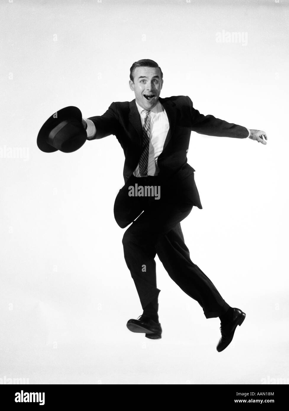 1960s MAN IN BUSINESS SUIT JUMPING INTO AIR WHILE RUNNING HAT IN HAND GOOFY FUNNY EXPRESSION ON HIS FACE HURRY SALESMAN WACKY Stock Photo
