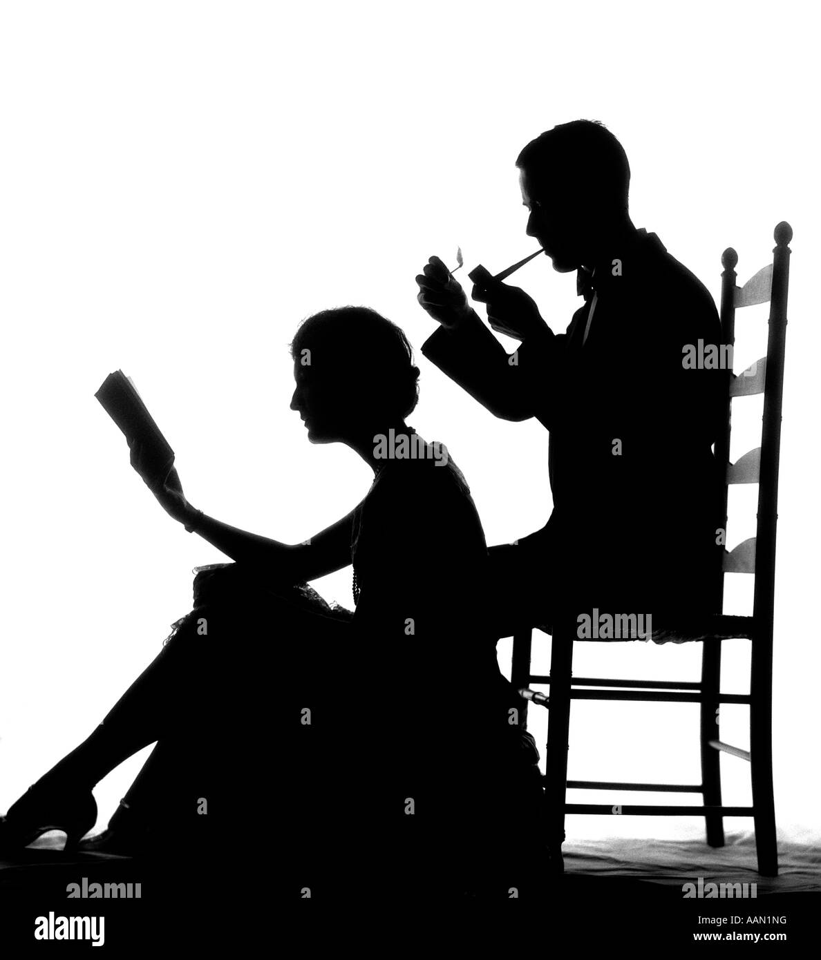 1920s 1930s ANONYMOUS SILHOUETTED MAN IN LADDER-BACK CHAIR SMOKING PIPE WITH WOMAN SEATED ON FLOOR IN FRONT OF HIM READING BOOK Stock Photo