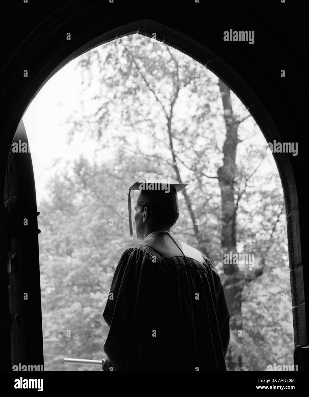 1960s BACK VIEW OF GRADUATE STANDING IN DARK ARCHED DOORWAY WITH TREES IN SUNLIT BACKGROUND Stock Photo