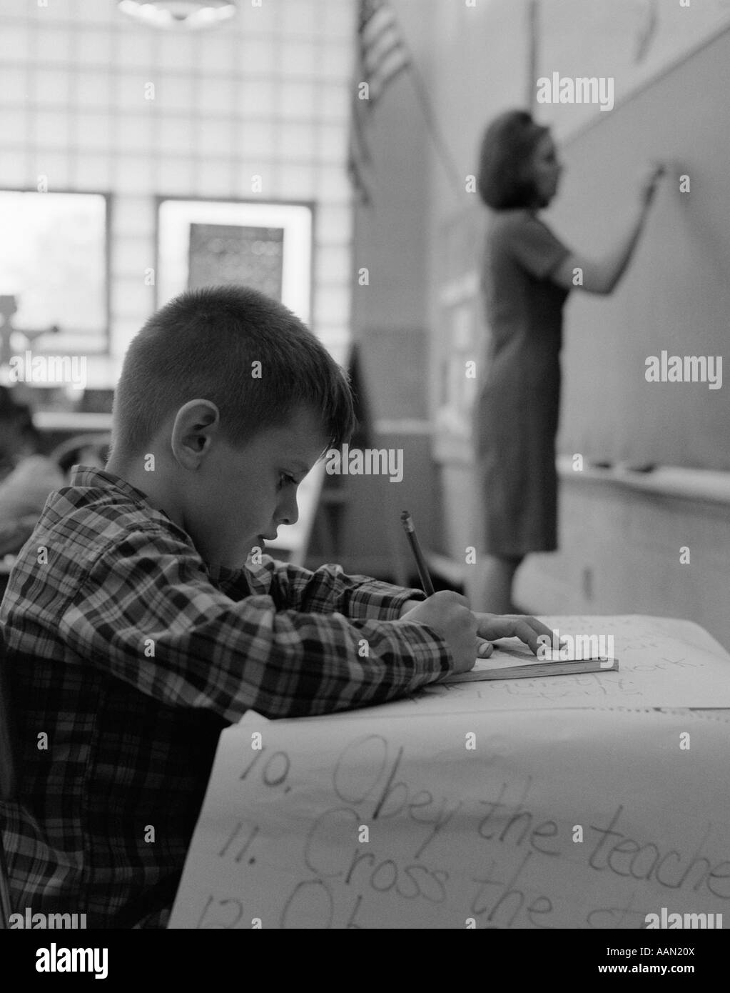 1960s SIDE VIEW OF GRADE SCHOOL BOY AT DESK WRITING WITH TEACHER WRITING ON BLACKBOARD IN SOFT FOCUS IN BACKGROUND Stock Photo