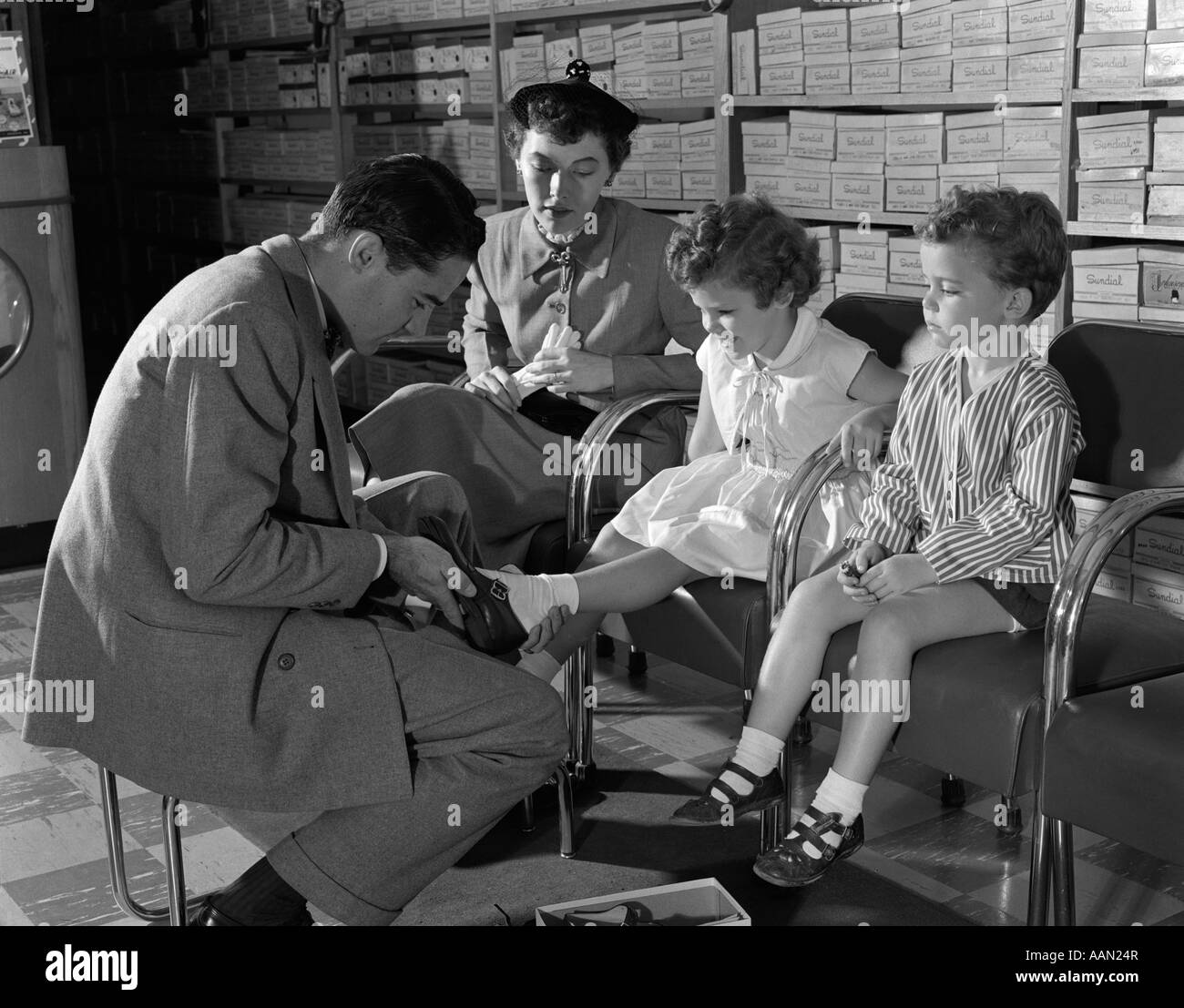1950s MOTHER SON DAUGHTER IN SHOE STORE SALESMAN SIZING SHOES ON LITTLE GIRL SHOP RETAIL SHELVES WITH SHOE BOXES Stock Photo