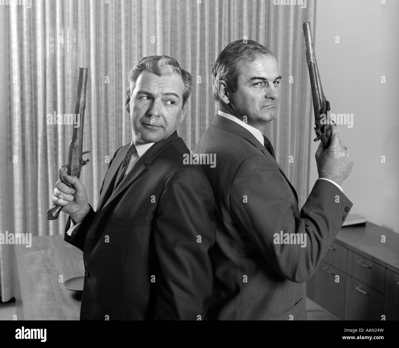 1960s 2 MIDDLE AGED MEN BACK TO BACK HOLDING DUEL PISTOLS GUNS Stock Photo