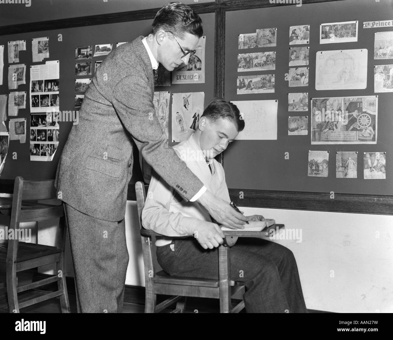 1940s STANDING MALE TEACHER INSTRUCTING SITTING MALE STUDENT IN FRONT OF BULLETIN BOARD CLASSROOM CONCENTRATION FACULTY Stock Photo