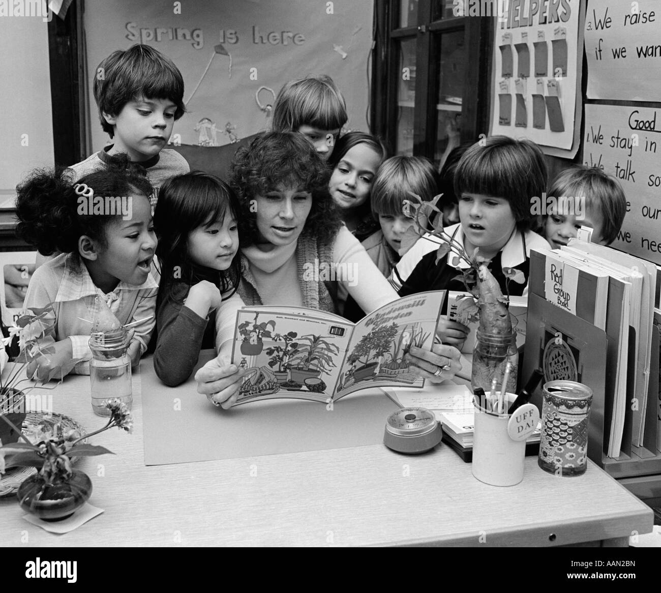 1980s TEACHER READING BOOK TO GROUP OF GRADE SCHOOL STUDENTS GATHERED AROUND HER Stock Photo