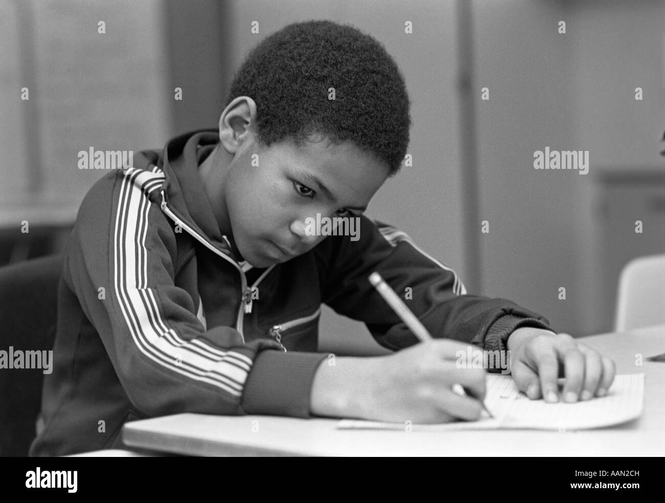 1980s AFRICAN AMERICAN MALE GRADE SCHOOL STUDENT SITTING AT DESK IN CLASSROOM WRITING Stock Photo