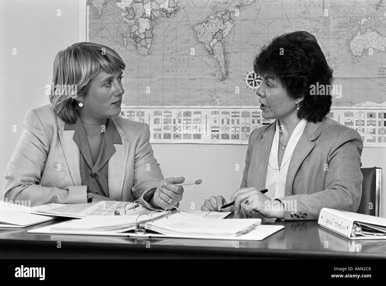 1980s PAIR OF FEMALE TEACHERS HAVING DISCUSSION NOTEBOOKS SPREAD OUT ON DESK IN FRONT OF THEM WORLD MAP ON WALL IN BACKGROUND Stock Photo