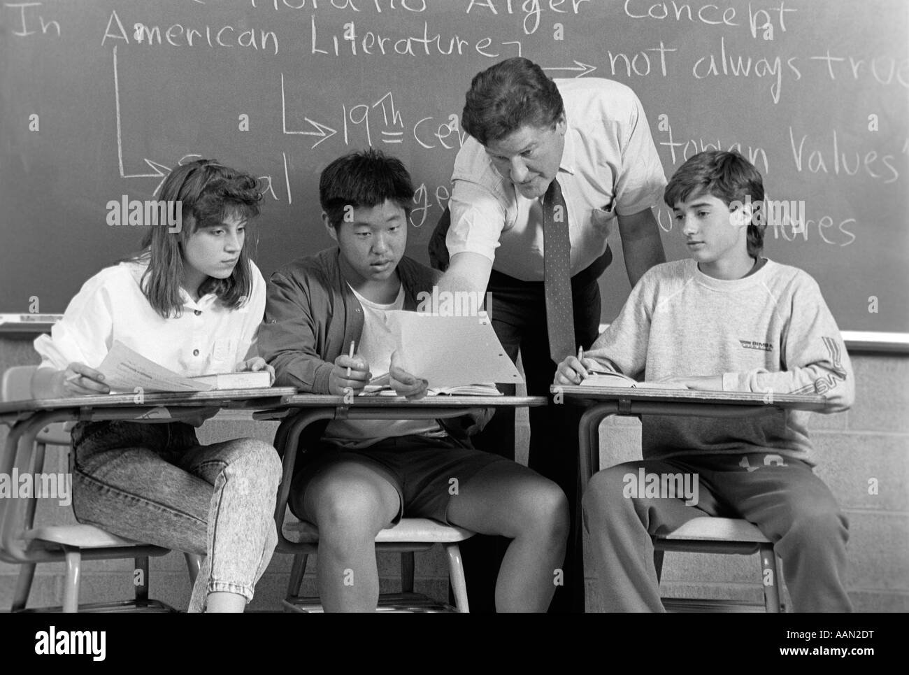 COLLEGE STUDENTS MEN WOMAN TEACHER IN CLASSROOM SITTING AT DESKS TEACHER LEANING OVER DESK EXAMINING PAPER OF STUDENT CHALKBOARD Stock Photo