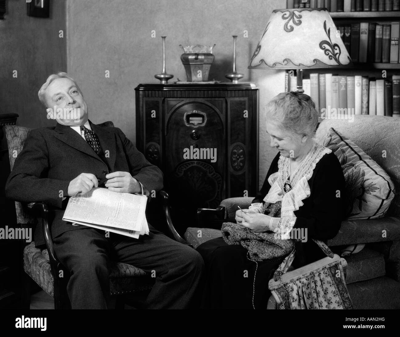 1930s ELDERLY COUPLE SITTING IN LIVING ROOM LISTENING TO RADIO WOMAN KNITTING MAN WITH NEWSPAPER IN LAP Stock Photo