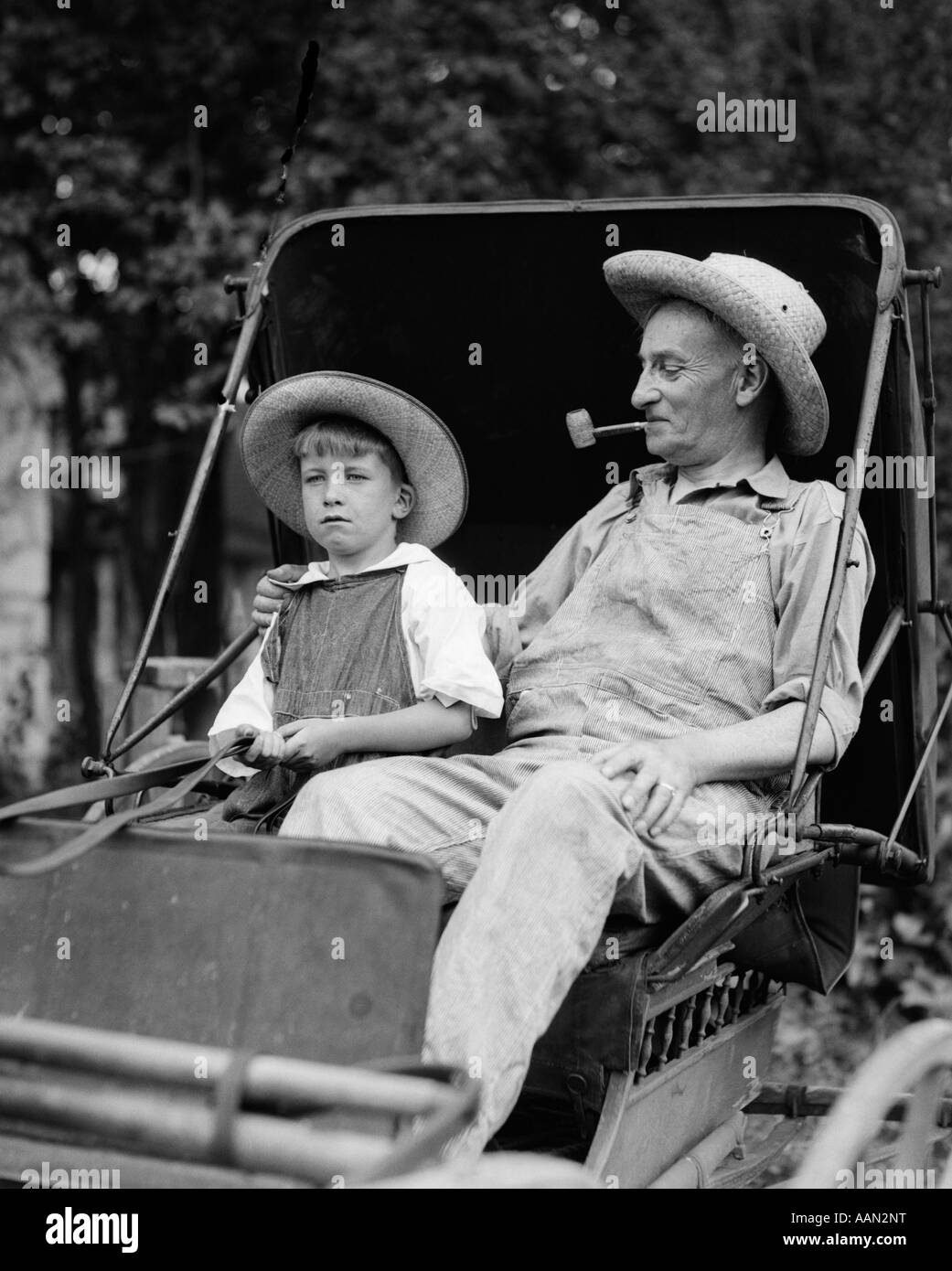 1930s FARM BOY & GRANDFATHER IN OVERALLS & STRAW HATS SITTING IN SMALL BUGGY Stock Photo