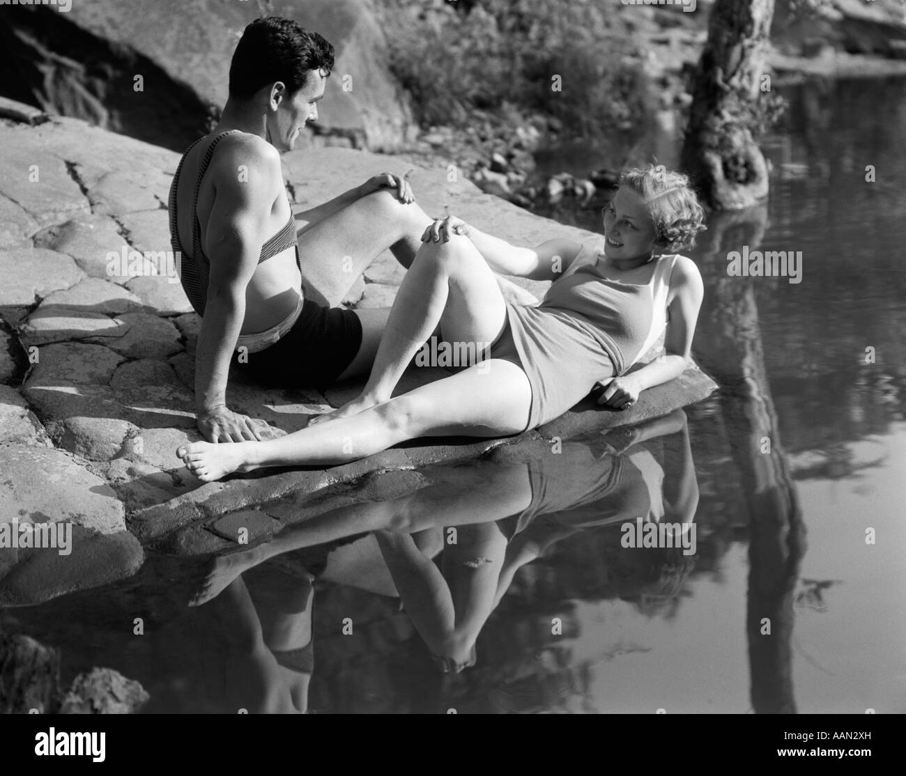 1930s COUPLE LYING ON ROCKS NEAR WATER WEARING BATHING SUITS Stock Photo