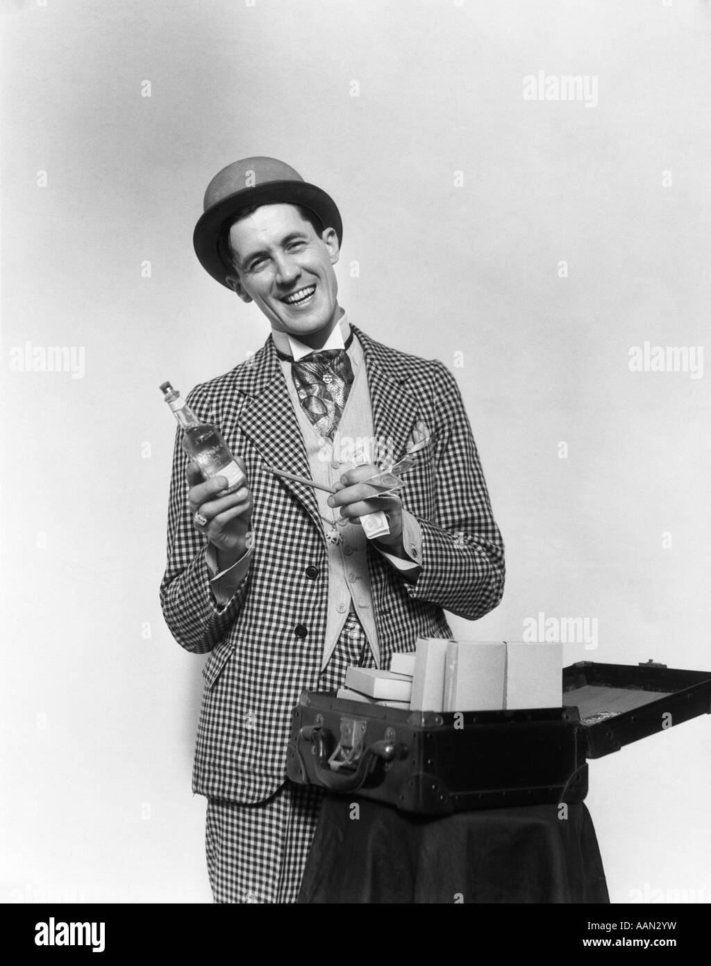1930s SMILING BARKER IN CHECKERED SUIT WITH MONEY IN HAND POINTING TO BOTTLE OF TONIC LOOKING AT CAMERA Stock Photo