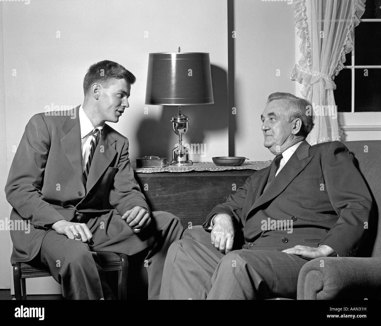 1950s YOUNG MAN SITTING IN LIVING ROOM TALKING TO SENIOR MAN Stock Photo