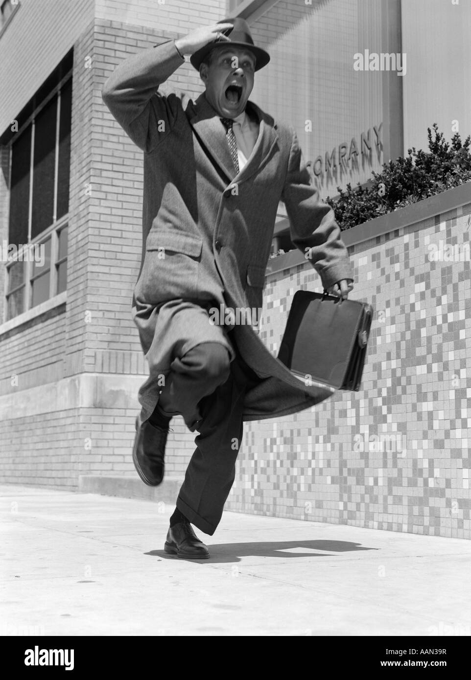 1960s 1950s FRANTIC MAN RUNNING DOWN STREET HOLDING HAT ON WITH HAND CARRYING BRIEFCASE WEARING TOP COAT Stock Photo