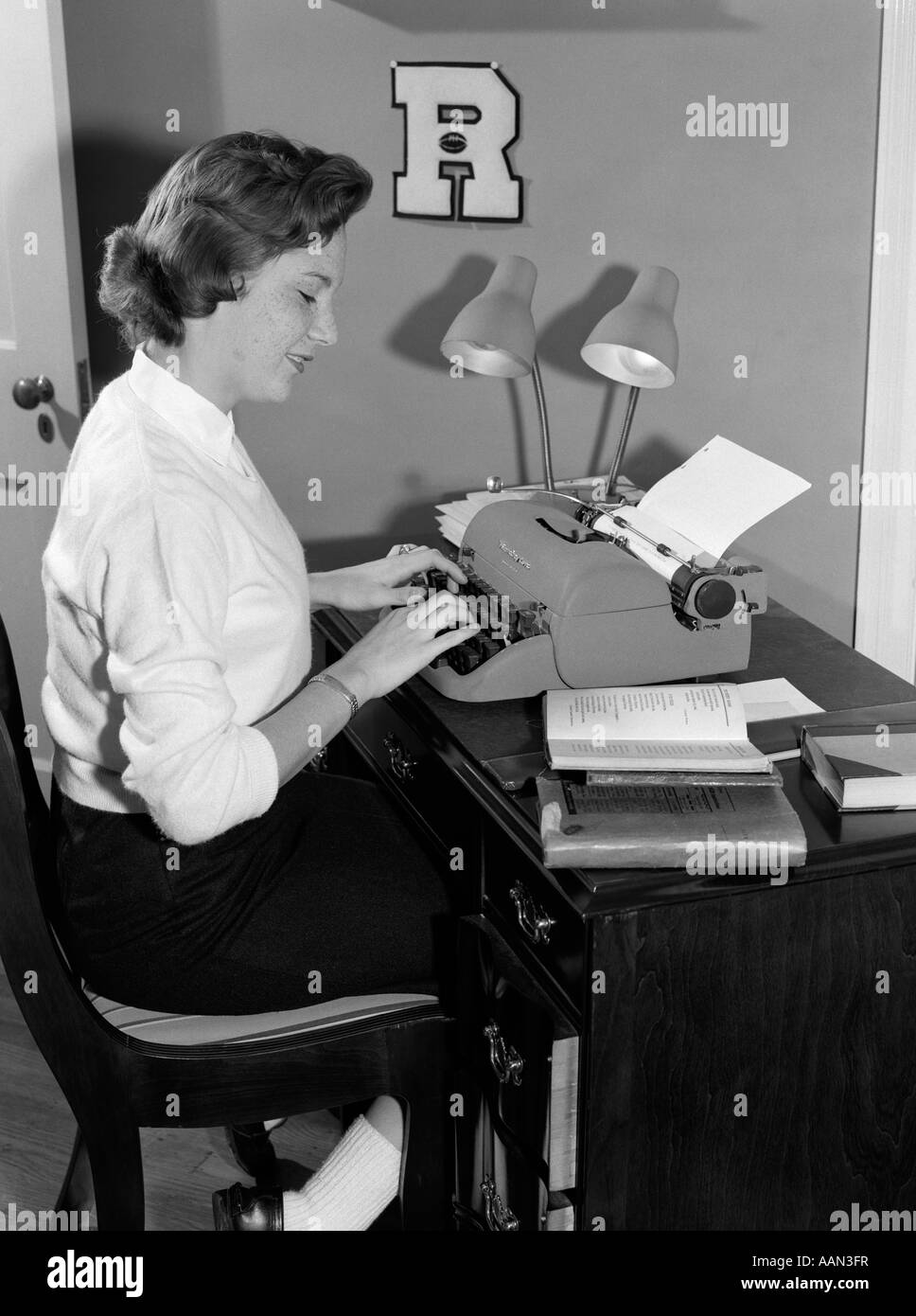 1950s FEMALE COLLEGE STUDENT SITTING AT DESK TYPING LOOKING AT BOOK BESIDE TYPEWRITER Stock Photo