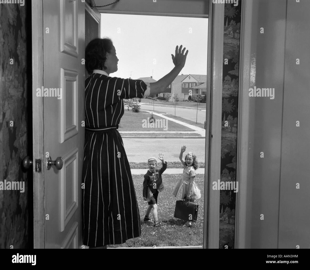 1950s WOMAN MOTHER WAVING FROM DOORWAY AS BOY SON AND GIRL DAUGHTER  LEAVE FOR SCHOOL IN MORNING Stock Photo