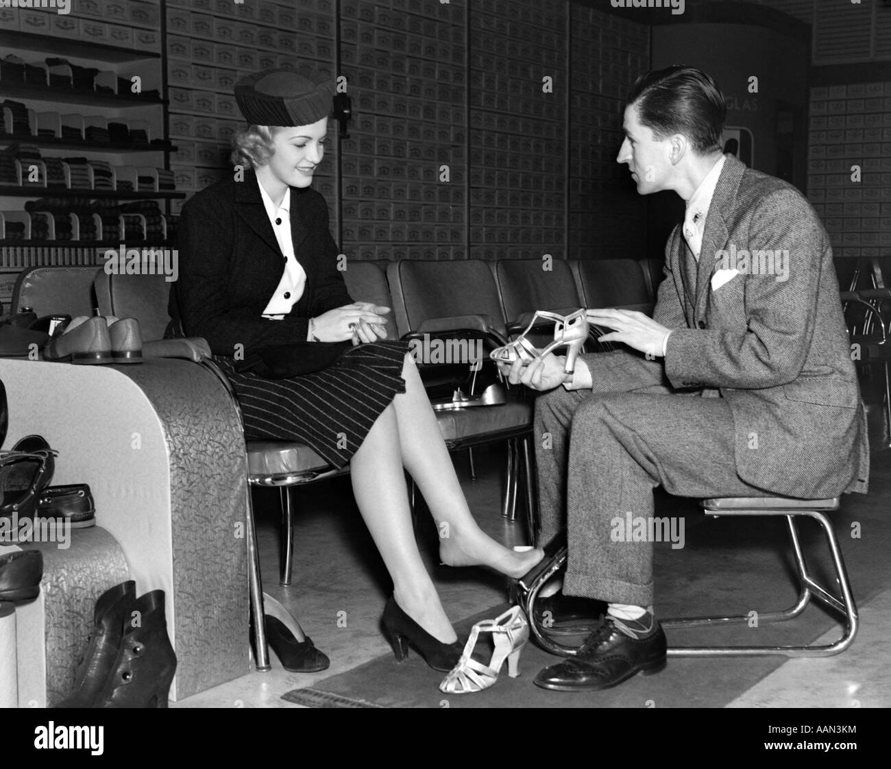 1930s SHOE SALESMAN HELPING WOMAN TRY ON PAIR HIGH HEELS IN RETAIL SHOE STORE Stock Photo