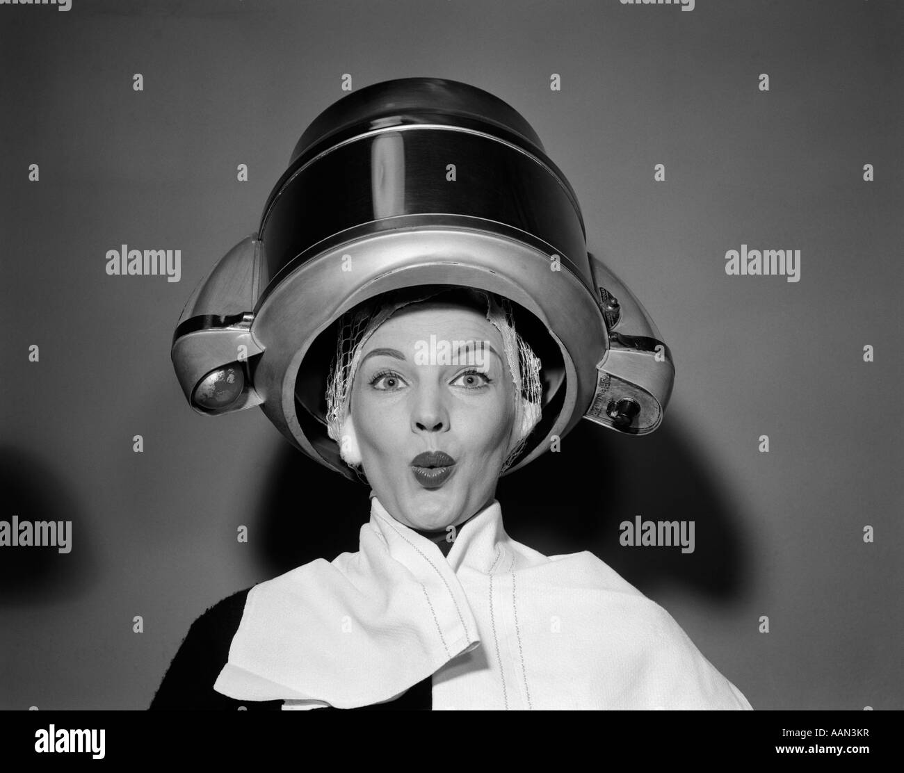 1950s WOMAN UNDER HAIR DRYER WITH TOWEL ON SHOULDERS AND HAIR NET LOOKING AT CAMERA Stock Photo
