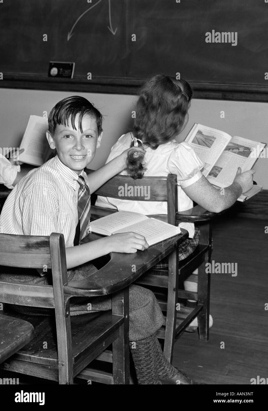 1940s 3 KIDS STUDENTS CLASSROOM BOYS GIRL FROM BACK ONE BOY TURNED SMILING AT CAMERA MISCHIEF OPEN BOOKS DESKS Stock Photo