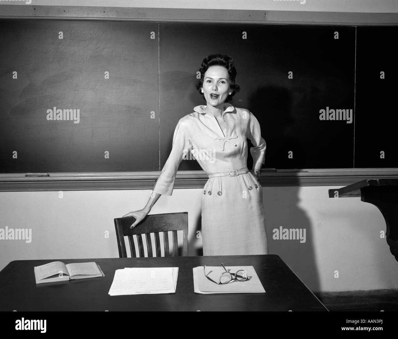 1950s SCHOOL TEACHER STANDING IN FRONT OF BLACKBOARD LEANING ON CHAIR BY DESK LOOKING AT CAMERA Stock Photo