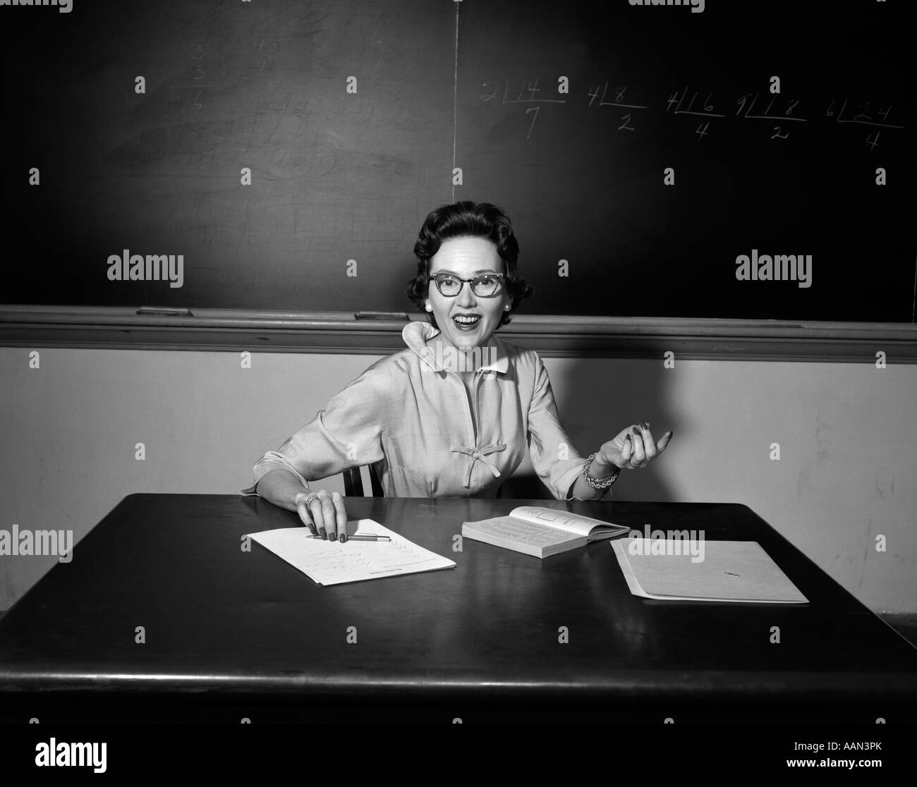 1950s SCHOOL TEACHER WEARING GLASSES SITTING AT DESK IN CLASSROOM LOOKING AT CAMERA Stock Photo