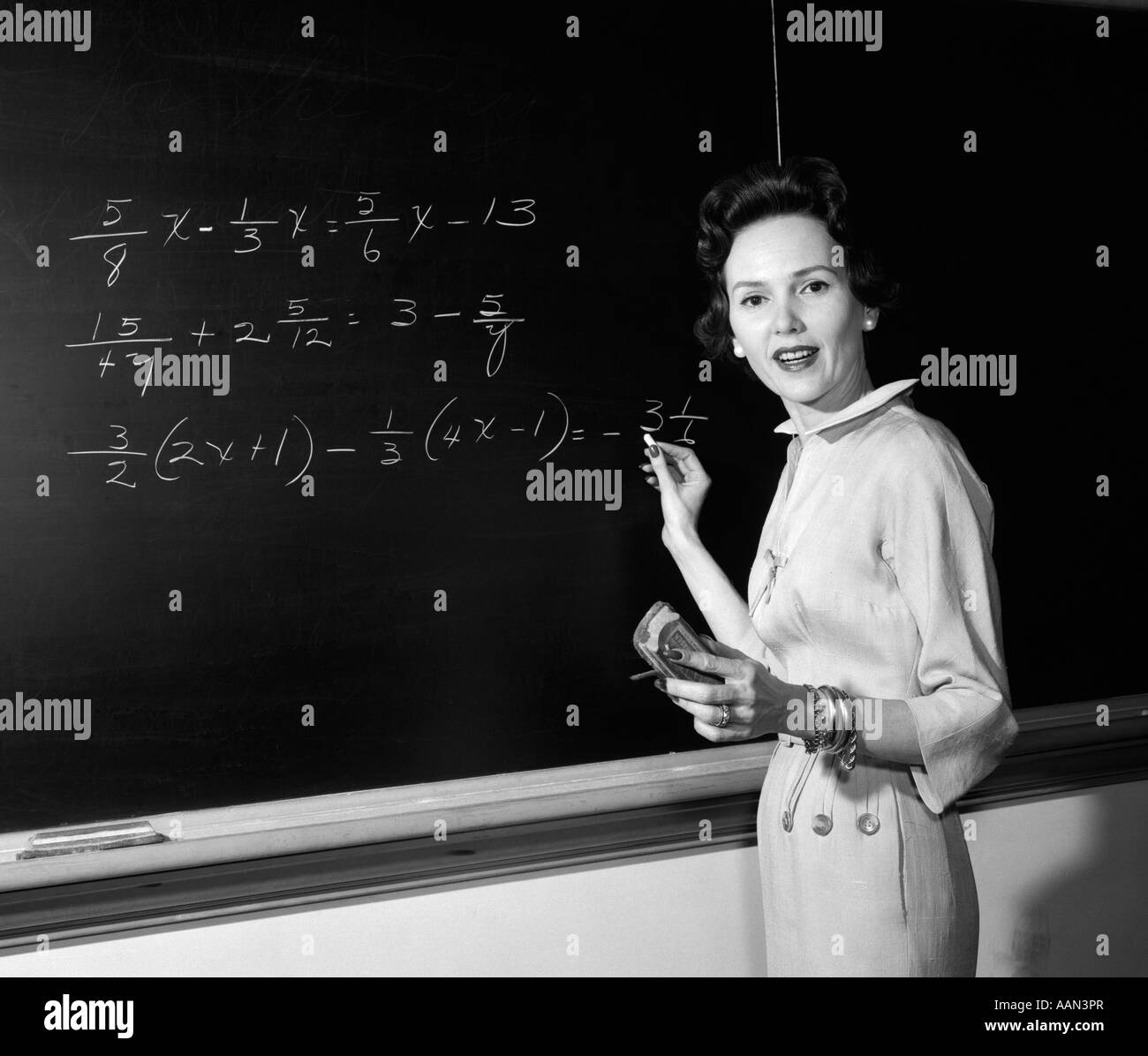 1950s MATH TEACHER STANDING IN FRONT OF BOARD HOLDING CHALK AND ERASER LOOKING AT CAMERA Stock Photo
