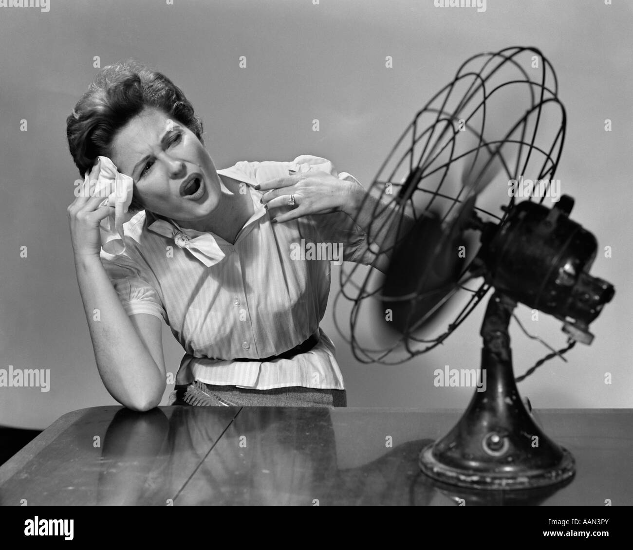 1950s VERY HOT WOMAN WIPING FOREHEAD SITTING IN FRONT OF FAN Stock Photo