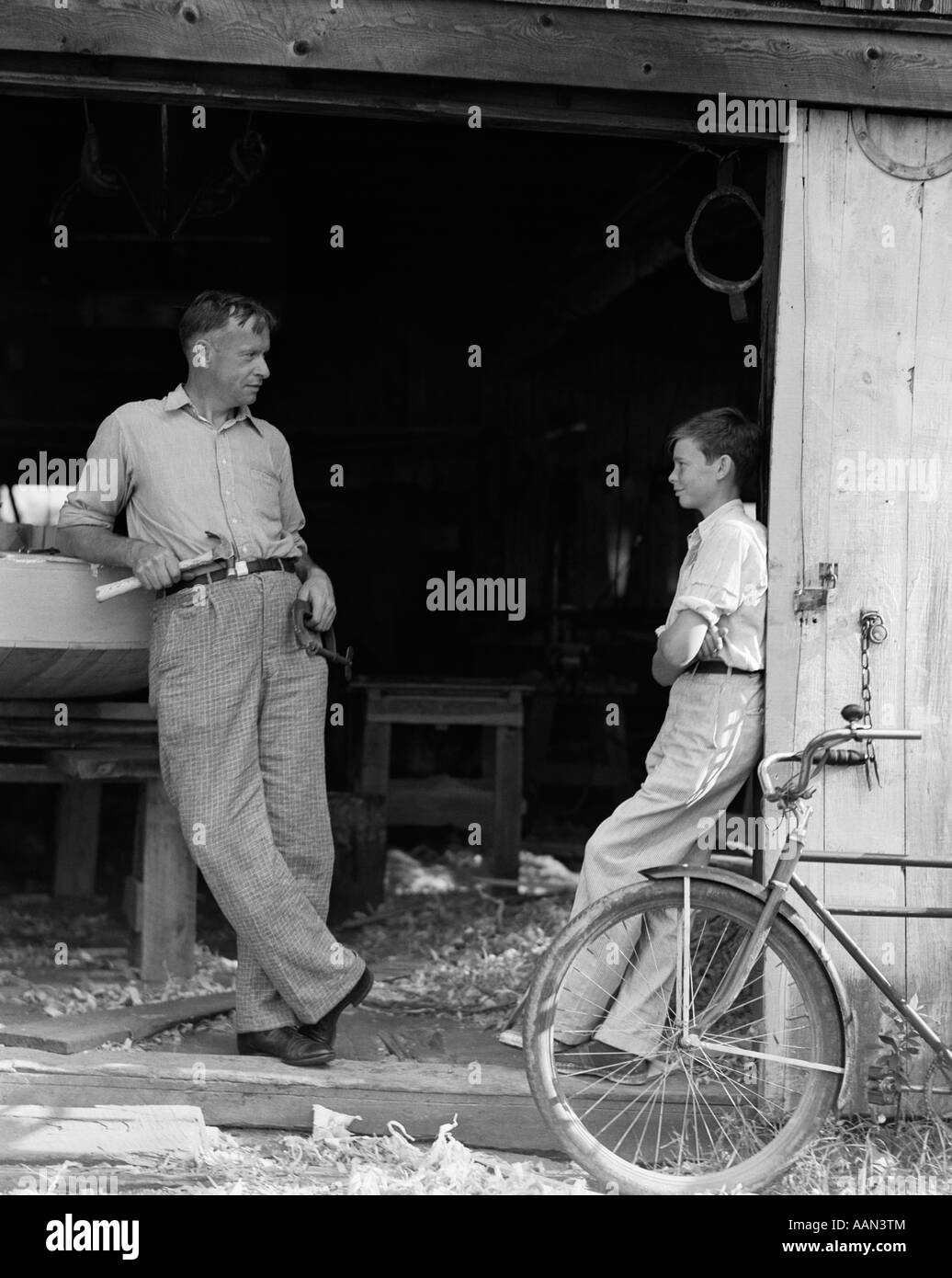 1930s MAN FATHER HOLDING HAND TOOLS TALKING TO BOY SON LEANING IN DOORWAY OF BOAT SHED Stock Photo