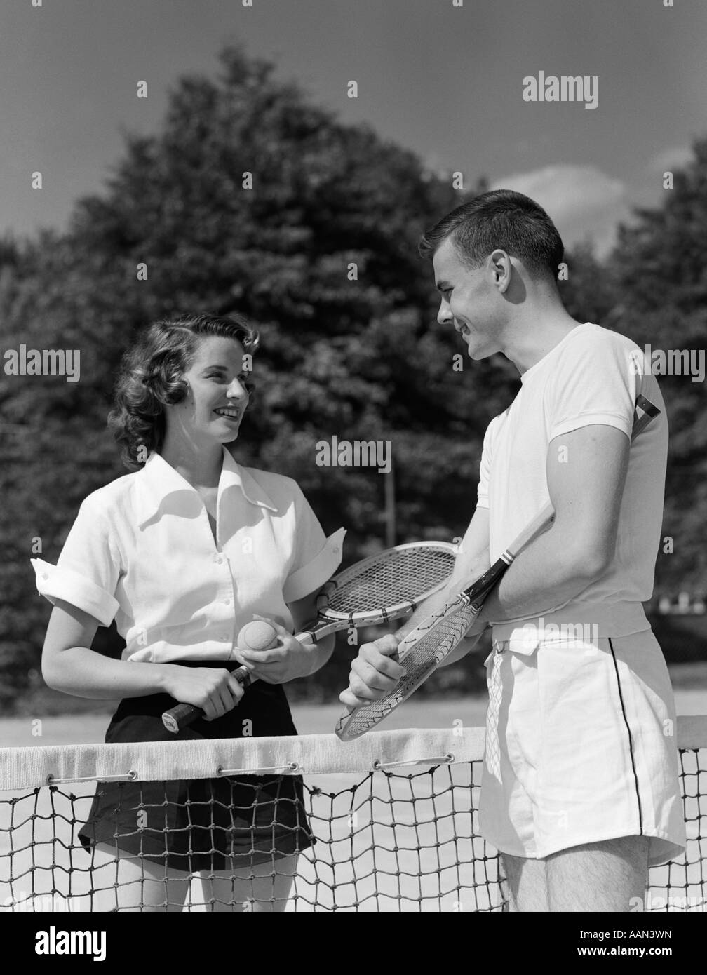 1950s COUPLE STANDING ON OPPOSITE SIDES OF NET HOLDING TENNIS RACQUETS Stock Photo