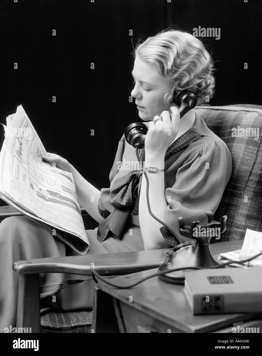 1920s 1930s YOUNG WOMAN SITTING IN CHAIR LOOKING AT NEWSPAPER TALKING ON TELEPHONE Stock Photo