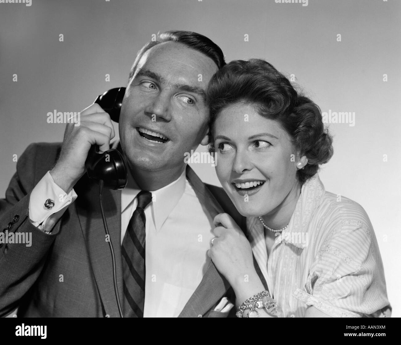 1950s 1960s MAN ON TELEPHONE WOMAN LEANING IN LISTENING BOTH SMILING Stock Photo
