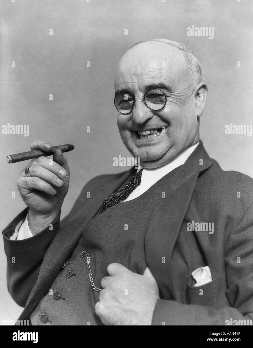 1930s ELDERLY BUSINESSMAN IN 3-PIECE SUIT & GLASSES LEANING BACK SMILING WITH CIGAR IN HAND Stock Photo