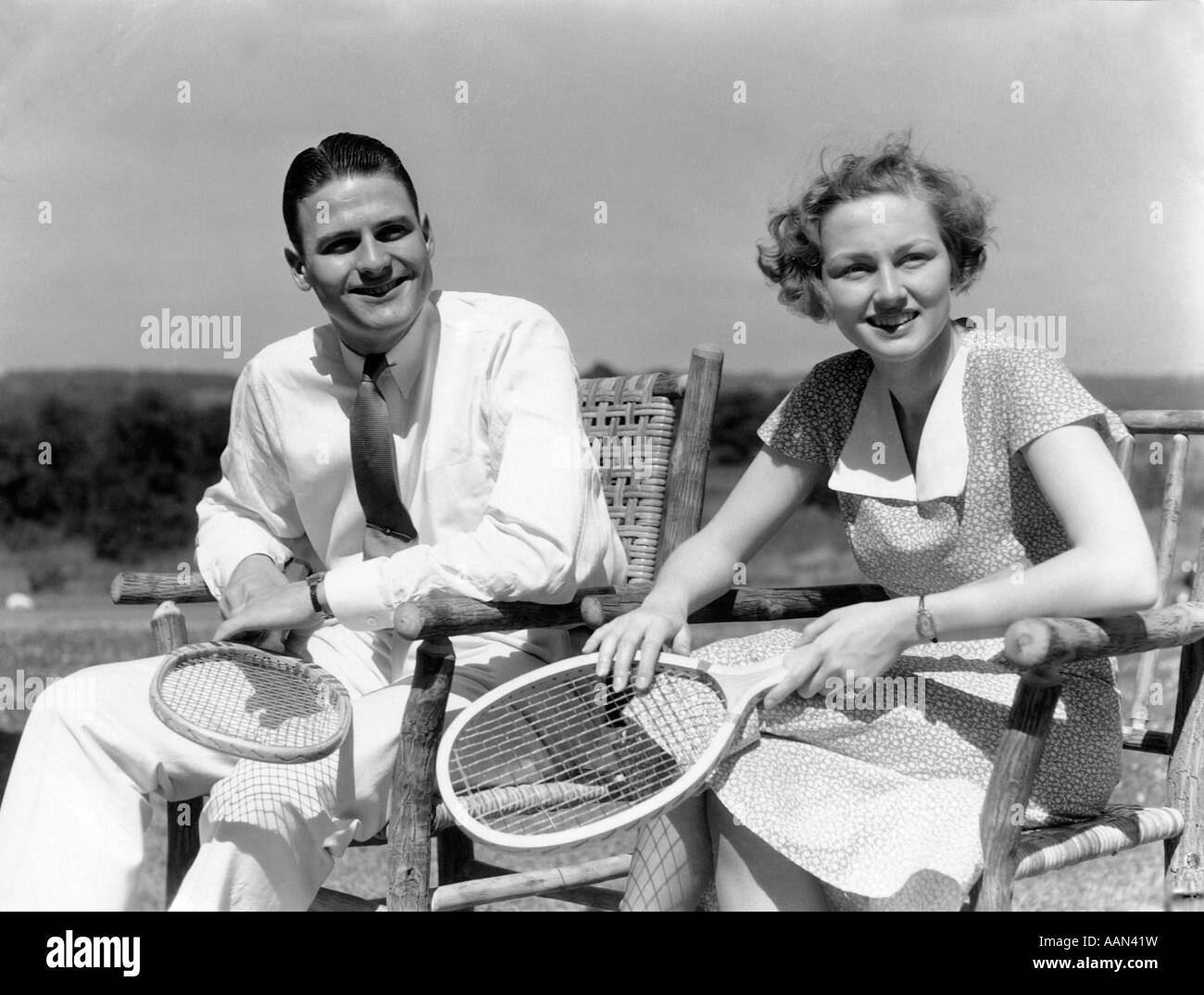 1940s MAN AND WOMAN SITTING IN WOODEN CHAIRS EACH HOLDING TENNIS RACQUET RACKET Stock Photo