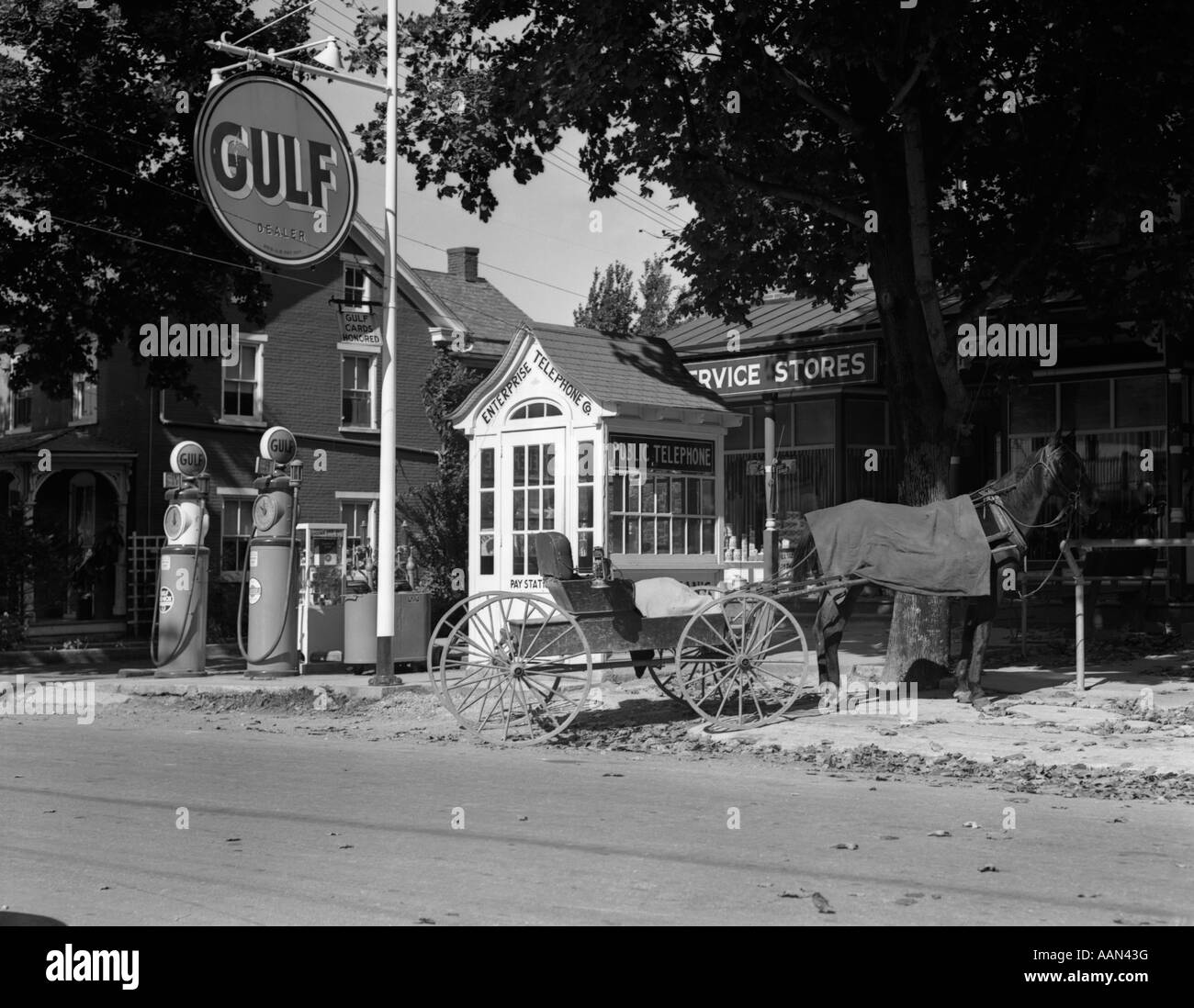 1930s HORSE & EMPTY BUGGY TIED TO POLE OUTSIDE OF GULF SERVICE STATION NEXT TO FANCY WOODEN PHONE BOOTH WITH SHINGLED ROOF Stock Photo