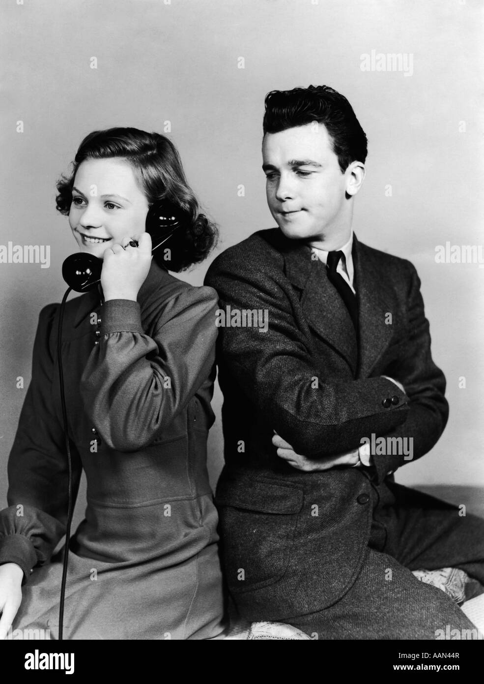 1940s YOUNG COUPLE GIRL TALKING ON TELEPHONE BOY ARMS CROSSED LOOKS ANNOYED Stock Photo