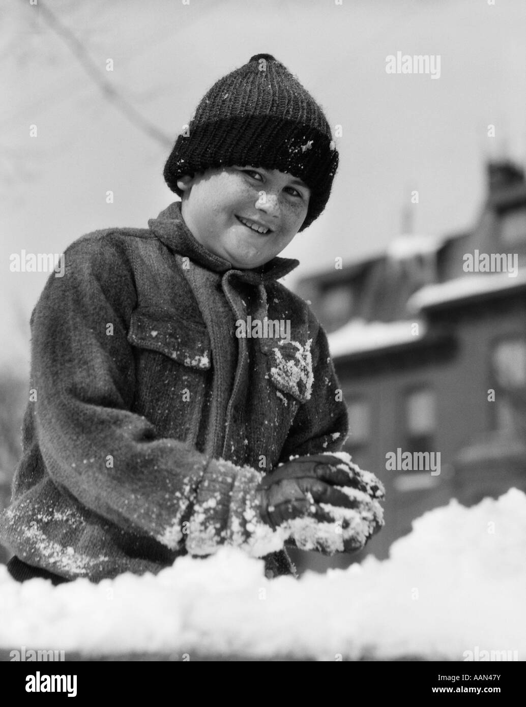 1920s 1930s SMILING BOY PLAYING IN SNOW MAKING SNOWBALL LOOKING AT CAMERA Stock Photo