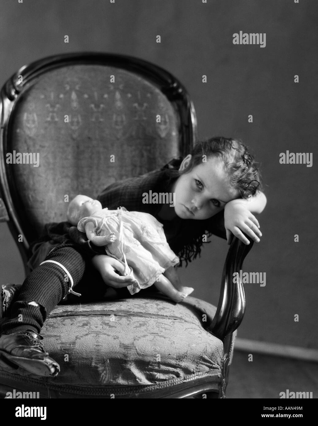 1930s LITTLE GIRL IN TATTERED CLOTHING WITH SAD EXPRESSION SITTING HOLDING DOLL LEANING HEAD ON WORN OUT CHAIR LOOKING AT CAMERA Stock Photo