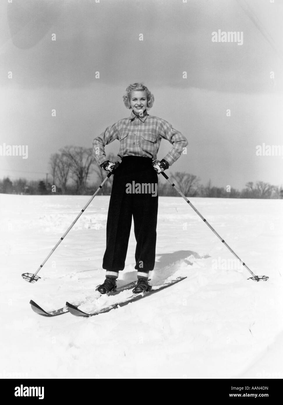 1940s SMILING BLOND WOMAN ON SKIS BOTH HANDS ON HIPS HOLDING SKI POLES IN SNOW FIELD Stock Photo