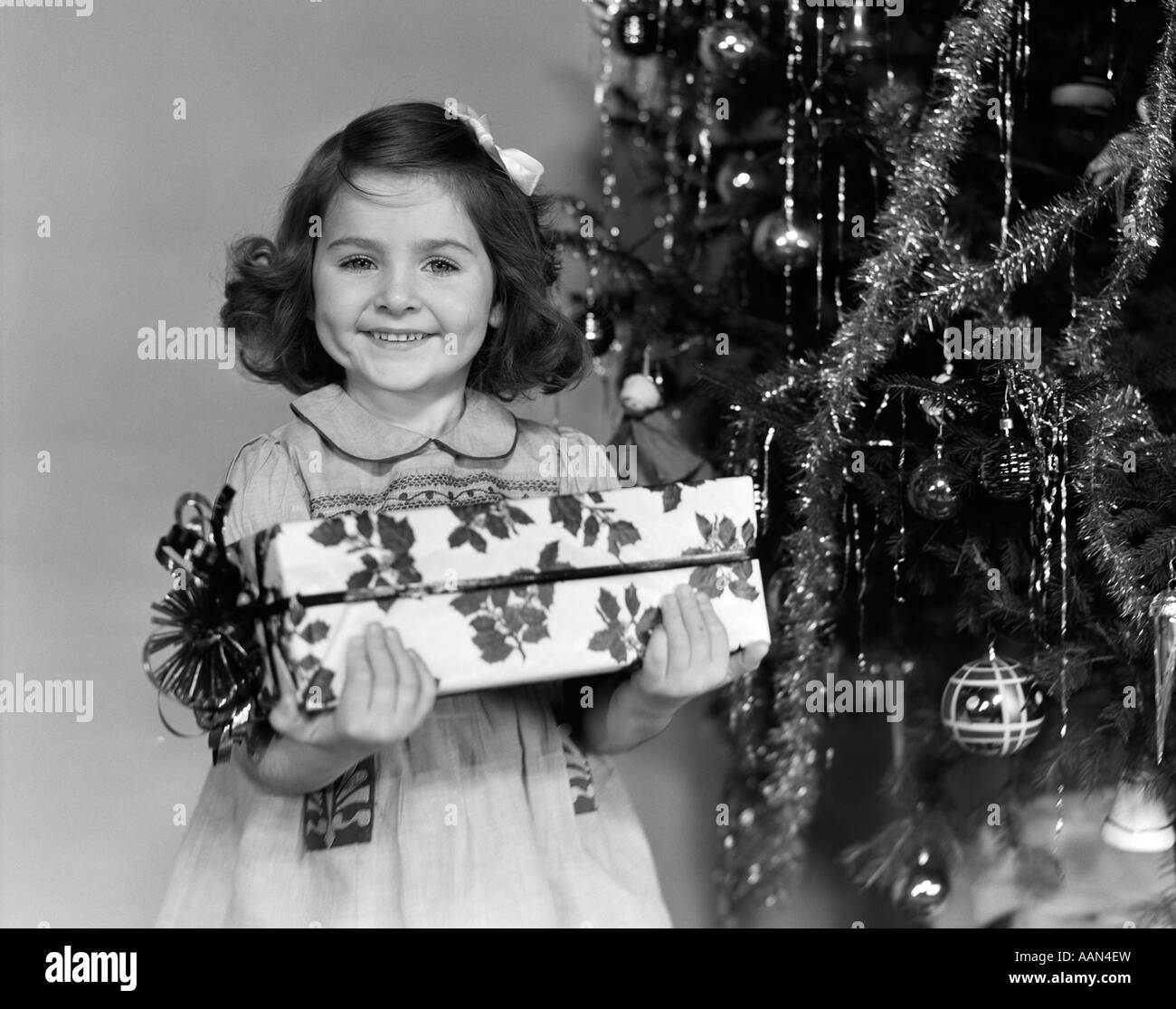 1940s LITTLE GIRL STANDING NEAR CHRISTMAS TREE HOLDING A WRAPPED PRESENT SMILING LOOKING AT CAMERA Stock Photo