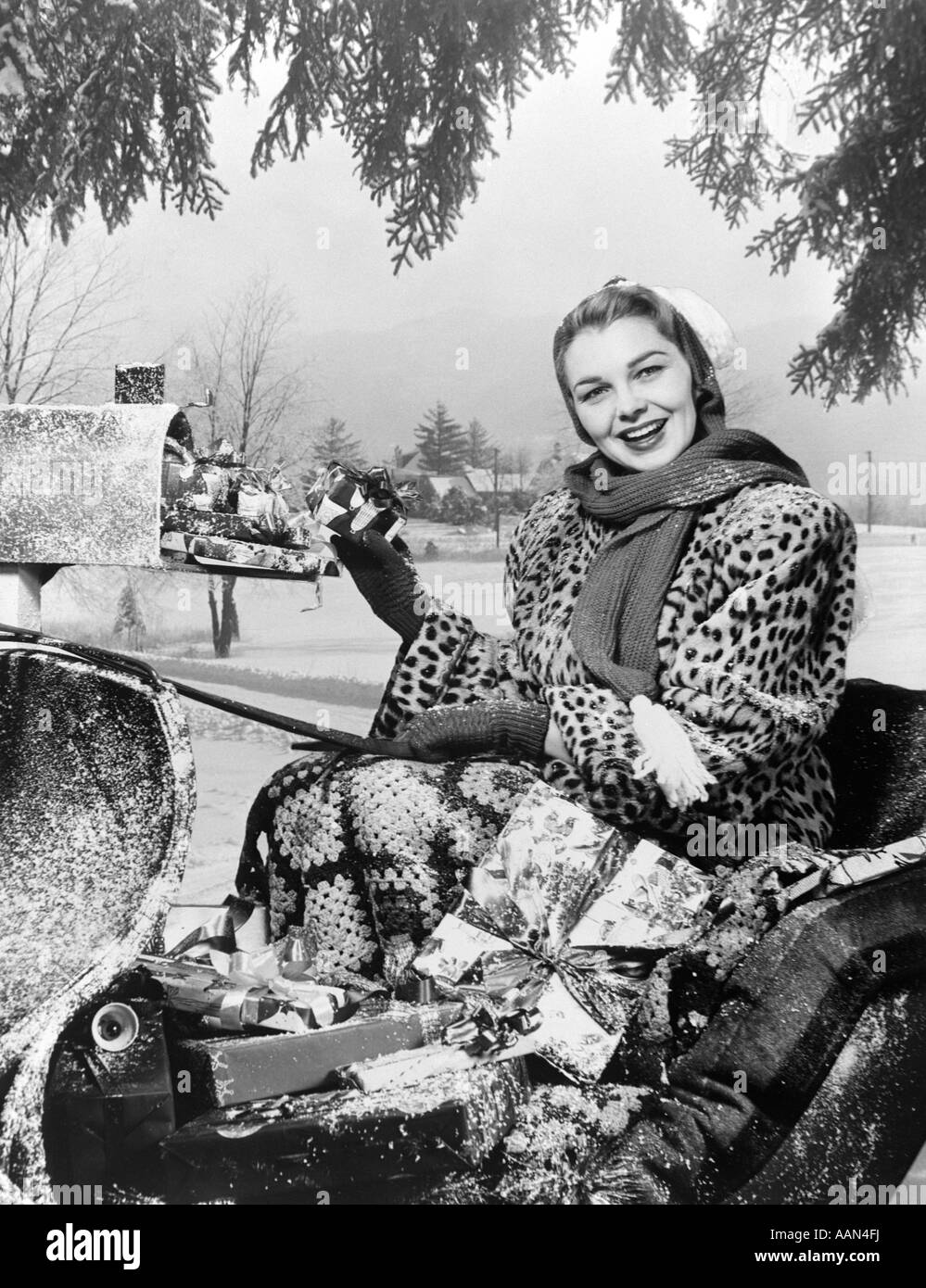 1950s SMILING WOMAN LOOKING AT CAMERA RIDING IN SLEIGH WEARING  LEOPARD SKIN FUR COAT AT MAILBOX WITH PACKAGES Stock Photo