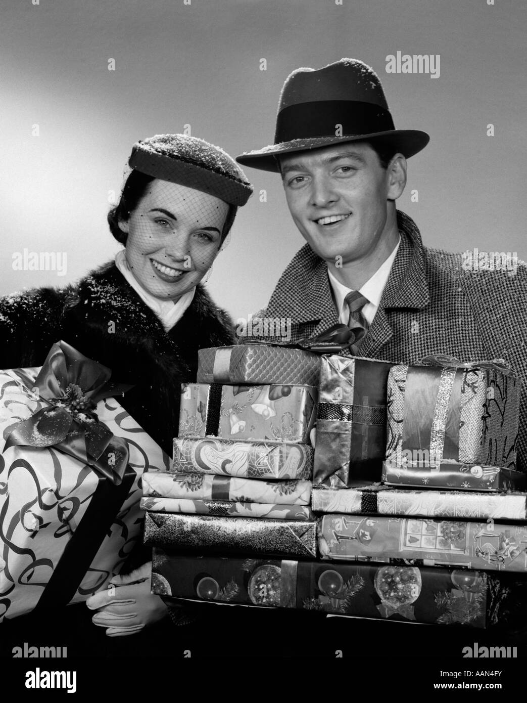 1950s 1960s SMILING PORTRAIT COUPLE IN WINTER COATS & HATS HOLDING WRAPPED CHRISTMAS GIFTS LOOKING AT CAMERA Stock Photo