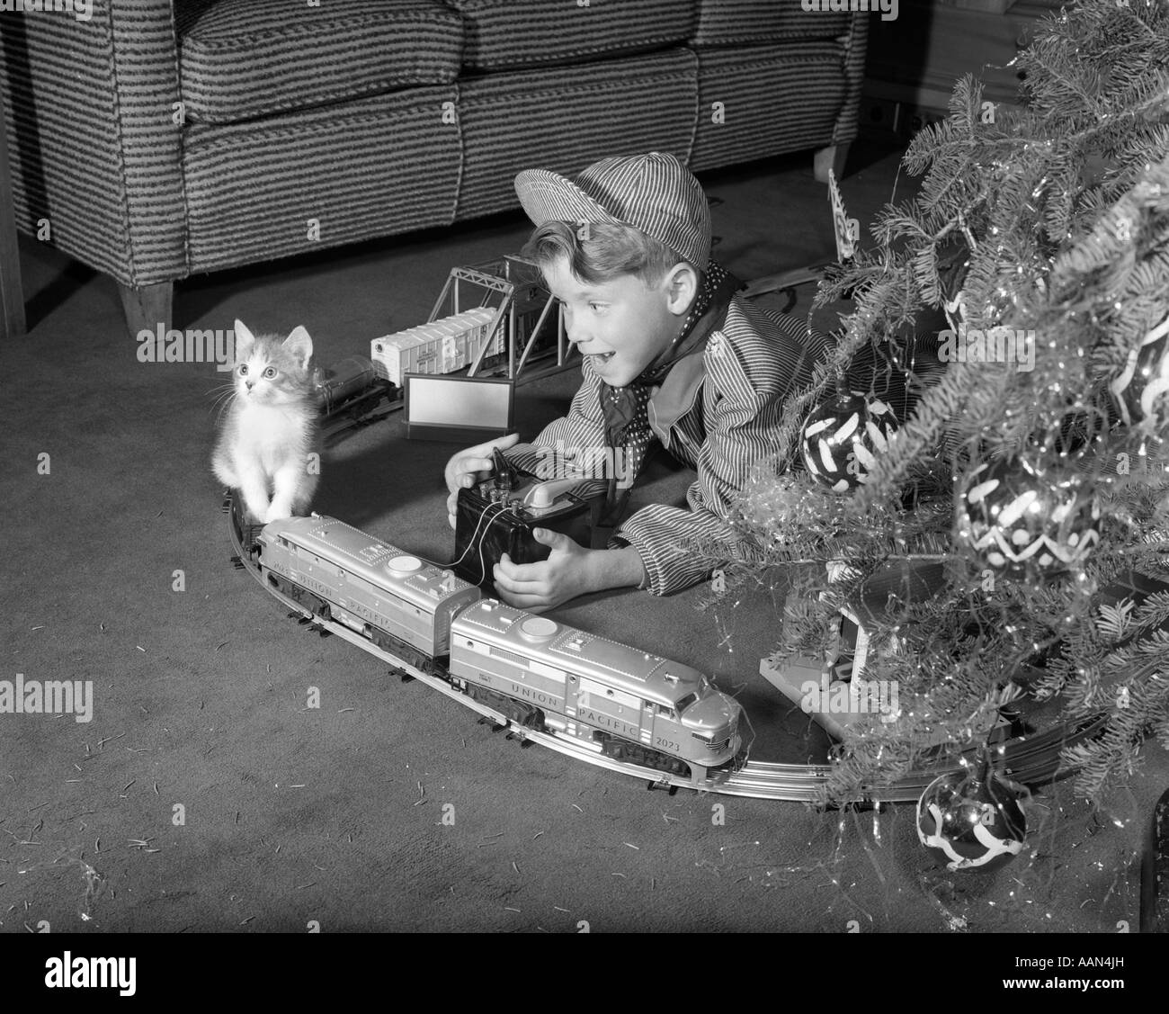 1950s BOY IN ENGINEER OUTFIT PLAYING WITH ELECTRIC TRAIN SET BY CHRISTMAS TREE AND KITTEN INDOOR Stock Photo